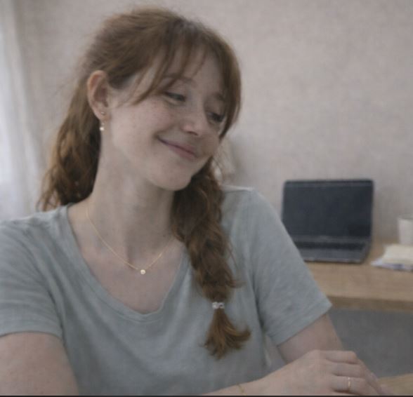 Jeune femme aux taches de rousseur assise à un bureau minimaliste, portant un collier coréen fin et des bijoux délicats, photo casual avec léger flou et lumière naturelle.