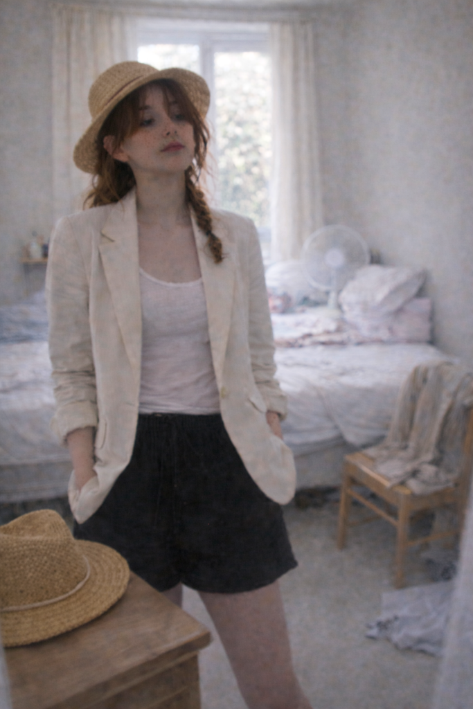 Jeune femme de 23 ans dans sa chambre, portant un blazer clair d’été, lumière surexposée, chapeaux de paille posés sur une table, scène du quotidien.