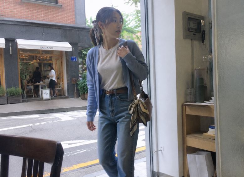 jeune femme asiatique portant un cardigan bleu ciel, un t-shirt écru et un jean avec un foulard imprimé noué à son sac dans un café à Séoul