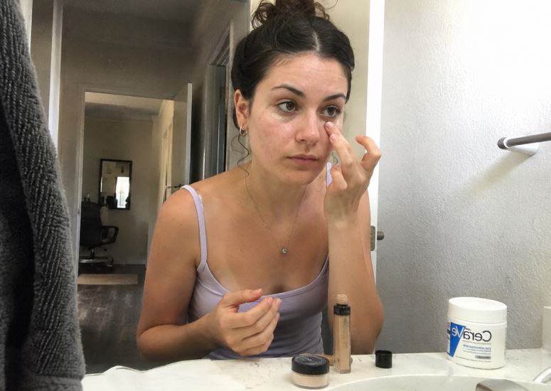 Jeune femme à peau claire appliquant du correcteur sous l’œil devant le miroir de sa salle de bain.