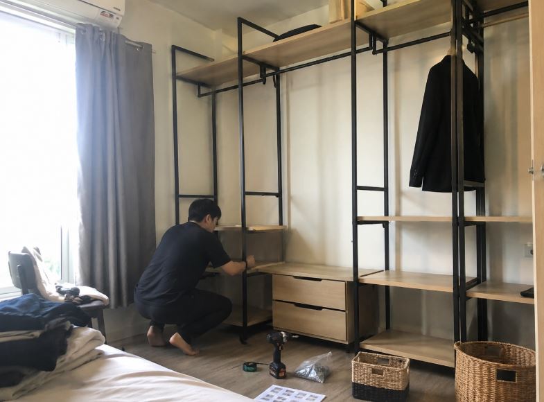 Jeune adulte asiatique installant une tablette en bois clair sur la structure noire d’un dressing modulable dans une petite chambre
