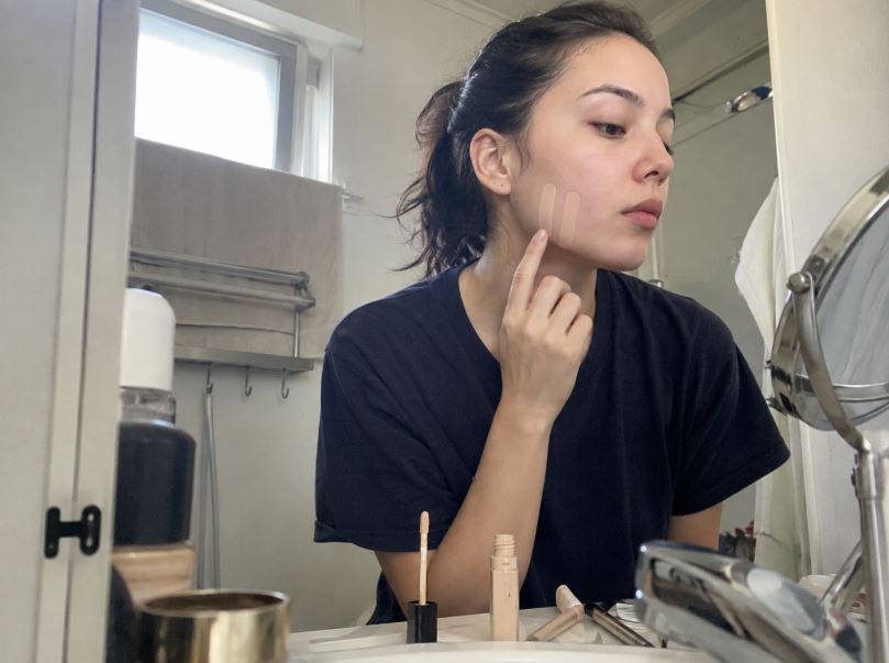 Jeune femme à peau claire testant deux nuances proches de correcteur beige clair sur la joue dans une salle de bain